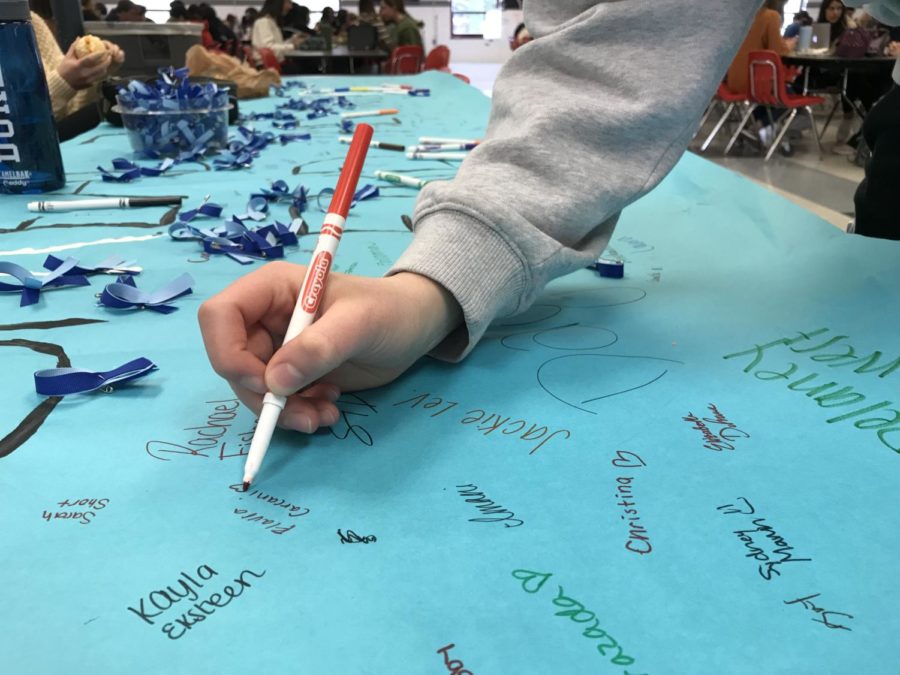 Junior Flavia Carcani signs the poster supporting the Spread the Word to End the Word campaign. The blue pins pictured here were handed out to all students in the cafeteria, as well. (Photo by Zach Anderson)